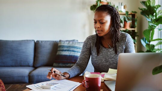 A woman sits at a table in front of an open laptop while using a pocket calculator.